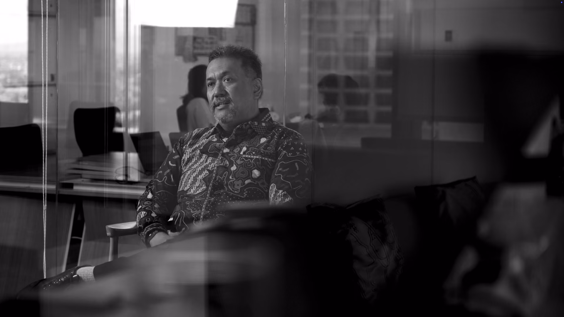 Black and white photo of a man wearing a patterned shirt sitting and talking in an office with glass walls and reflections.