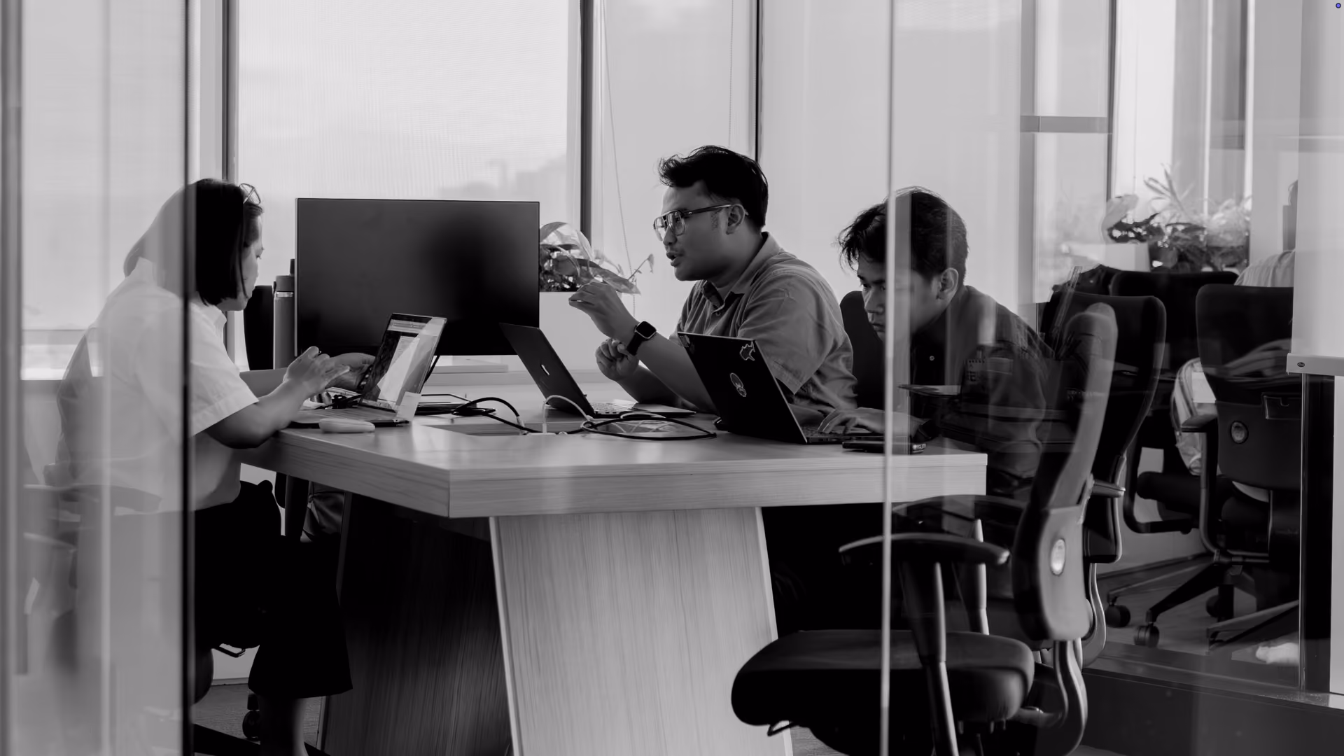 Three people sitting at a conference table in a glass-walled office, each working on laptops and engaged in discussion.