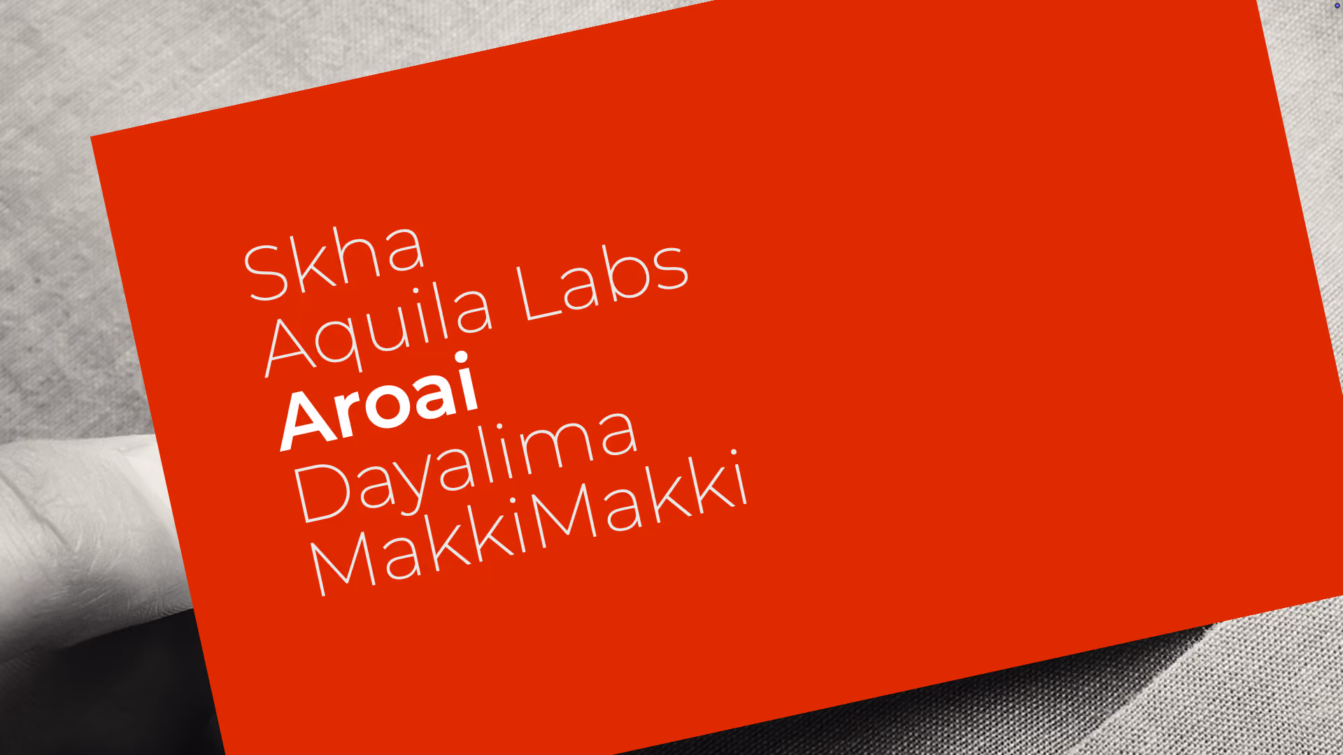 A hand holding a red card with white text listing names: Skha, Aquila Labs, Aroai in bold, Dayalima, and MakkiMakki.
