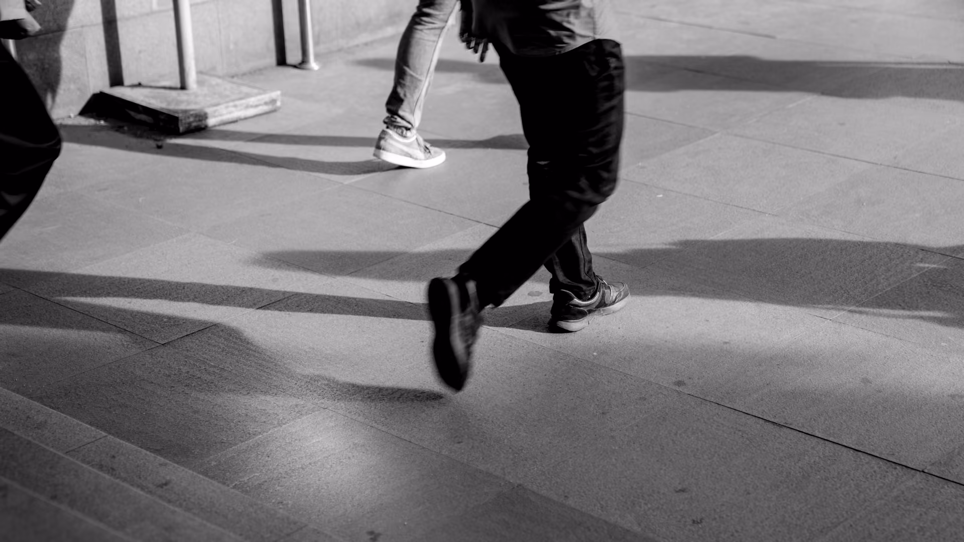 Black and white photo showing legs and feet of people walking on a paved surface next to steps, with noticeable shadows cast on the ground.